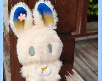May include: A plush bunny toy with cream-colored fur, blue and yellow accents on the ears, and light blue eyes. A small pink flower is on one ear. The bunny has a small white belly with a yellow design.