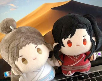 Può includere: Due bambole di peluche con tratti del viso ricamati. Una bambola è vestita di bianco e azzurro, l'altra di rosso. Entrambe le bambole hanno i capelli neri e sono sedute sulla tastiera di un laptop.