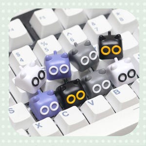 May include: A collection of custom keycaps in various colors, including white, gray, black, and purple, each featuring a cartoon cat face design with large, round eyes. The keycaps are arranged on a white keyboard.