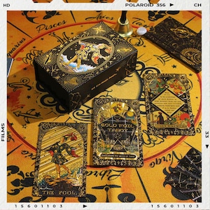 May include: A black and gold tarot card box and cards are displayed on a yellow cloth with astrological symbols. The box and cards feature gold foil accents and illustrations. The text "GOLD FOIL TAROT" is visible on one card, and "THE FOOL" on another.