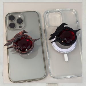 Puede incluir: Dos fundas transparentes para tel&eacute;fono con adornos decorativos. Una funda tiene un cuervo color bronce sobre una gema roja facetada. La otra funda tiene un cuervo negro sobre una gema similar. Sobre un fondo claro.