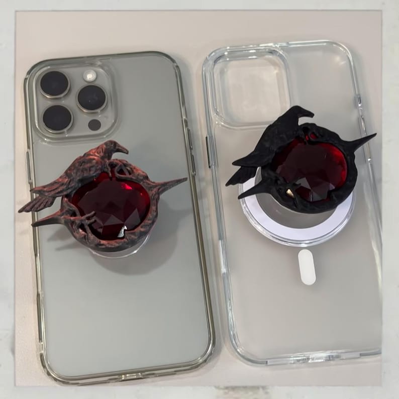 Puede incluir: Dos fundas transparentes para tel&eacute;fono con adornos decorativos. Una funda tiene un cuervo color bronce sobre una gema roja facetada. La otra funda tiene un cuervo negro sobre una gema similar. Sobre un fondo claro.