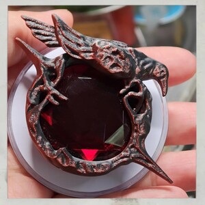 Puede incluir: Un colgante detallado, rojo oscuro y negro, con una gran gema facetada de color rub&iacute;. El colgante tiene un dise&ntilde;o ornamental, similar a un p&aacute;jaro, con detalles intrincados y est&aacute; sobre un fondo blanco.