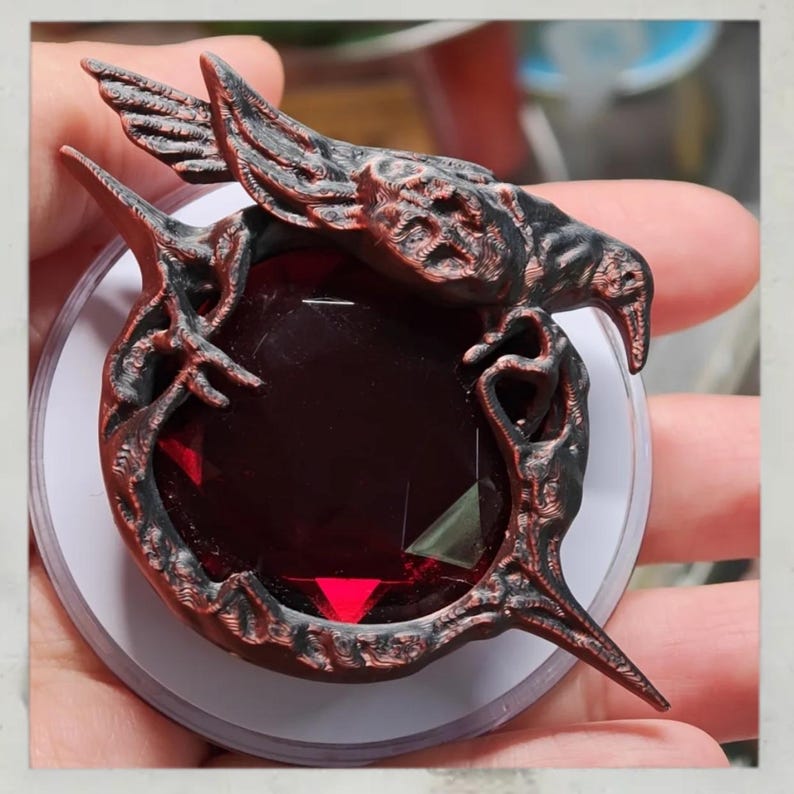Puede incluir: Un colgante detallado, rojo oscuro y negro, con una gran gema facetada de color rub&iacute;. El colgante tiene un dise&ntilde;o ornamental, similar a un p&aacute;jaro, con detalles intrincados y est&aacute; sobre un fondo blanco.