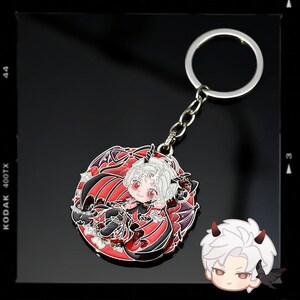 May include: A silver keychain with a detailed, colorful charm. The charm features a cartoon character with white hair, red eyes, and horns, surrounded by red and black wings. The design is set against a red background.
