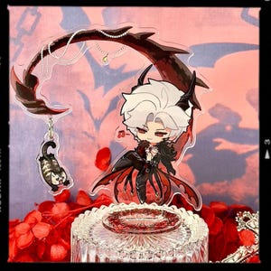 May include: A clear acrylic charm featuring a chibi character with white hair and red eyes, dressed in black and red attire. A black cat charm hangs below. The charm is displayed on a glass stand with red accents.