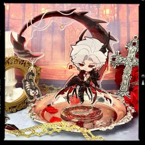 May include: A decorative acrylic figure of a character with white hair and red eyes, wearing a black and red outfit. The figure is on a silver tray with red rose petals. A matching keychain of a cat is also present.