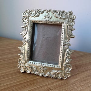 May include: A rectangular picture frame with a metallic gold finish. The frame features an ornate, raised relief border with scrolling leaf patterns and a fleur-de-lis motif at the top center. A row of small beads lines the inner edge of the frame.