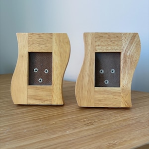 May include: Two light-colored wooden picture frames with a wavy, curved side profile. Each frame features a rectangular center opening with a dark brown backing and three small, circular metal fasteners arranged in a triangular pattern.