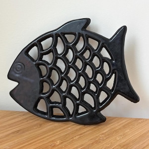 Könnte beinhalten: Ein schwarzer, fischförmiger Untersetzer mit einem ausgeschnittenen, durchbrochenen Design, das Fischschuppen nachahmt. Das Objekt hat eine glatte, glänzende Oberfläche und einen massiven Kopfbereich mit einem kreisförmigen Augendetail.