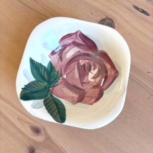 May include: A square, off-white ceramic dish with a hand-painted rose design. The rose is a deep burgundy color with green leaves. The dish has a slightly irregular shape and a glossy finish.