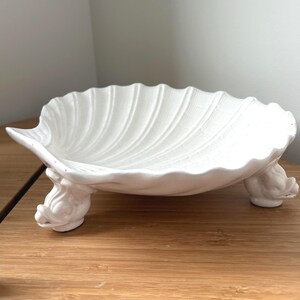 Puede incluir: Un plato de cerámica blanca con forma de concha de vieira, que presenta una superficie interior acanalada y se apoya sobre tres pequeños pies esculpidos que parecen cabezas estilizadas de delfines o criaturas marinas.