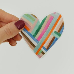 May include: A heart-shaped sticker with a colorful, geometric design. The design features a series of intersecting lines in various colors, including pink, blue, green, orange, and brown. The sticker is held against a white background.