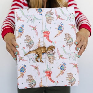 May include: A white gift wrap with a repeating pattern of dinosaurs wearing sweaters. A gold dinosaur toy is attached to the wrapping paper with twine.