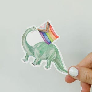 May include: A green dinosaur sticker with a rainbow flag held in its mouth. The sticker is held by a hand with white nail polish against a white background. The dinosaur is a watercolor illustration.