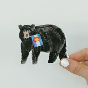 May include: A sticker featuring a black bear with a Colorado state flag. The bear is depicted in a watercolor style, with a detailed fur texture. The flag is attached to a small stick held in the bear's mouth.