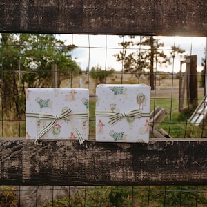May include: Two wrapped gifts with sheep and festive illustrations, tied with striped ribbon bows. The gifts rest on a weathered wooden fence, with a wire mesh fence and a natural landscape in the background.
