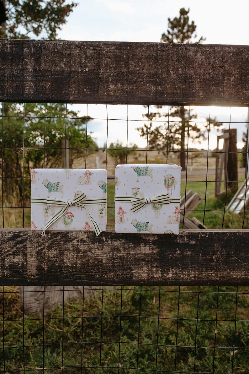 May include: Two wrapped gifts with sheep and festive illustrations, tied with striped ribbon bows. The gifts rest on a weathered wooden fence, with a wire mesh fence and a natural landscape in the background.