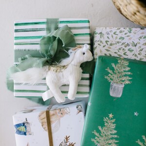 Puede incluir: Tres regalos envueltos con diferentes dise&ntilde;os. El regalo de arriba est&aacute; envuelto en rayas verdes y blancas con un adorno de unicornio de peluche blanco. El regalo del medio est&aacute; envuelto en blanco con una cinta marr&oacute;n y un patr&oacute;n de gatos con su&eacute;teres. El regalo de abajo est&aacute; envuelto en verde con un patr&oacute;n de &aacute;rboles de Navidad en macetas.