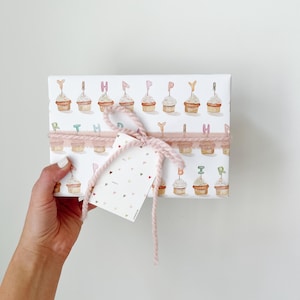 May include: A wrapped gift box with a cupcake pattern and the word "HAPPY" spelled out on top. The box is tied with pink yarn and a small tag. The gift is held by a person's hand.