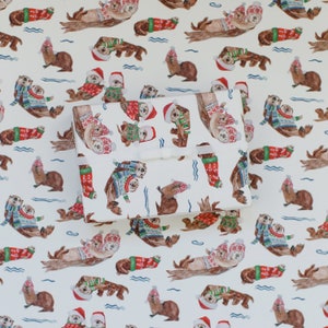 May include: White wrapping paper with a repeating pattern of sea otters wearing Christmas sweaters. The otters are in various poses, some swimming, some holding paws, and some holding fish. The sweaters are red and green with white snowflakes and a white trim.