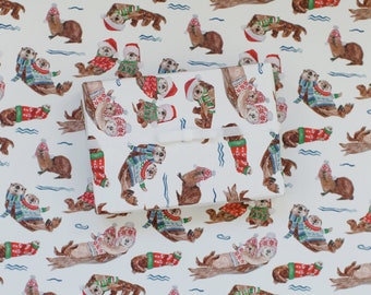 Könnte beinhalten: Weißes Geschenkpapier mit einem sich wiederholenden Muster von Seeottern, die Weihnachtspullover tragen. Die Otter sind in verschiedenen Posen, einige schwimmen, einige halten Pfoten und einige halten Fische. Die Pullover sind rot und grün mit weißen Schneeflocken und einem weißen Besatz.