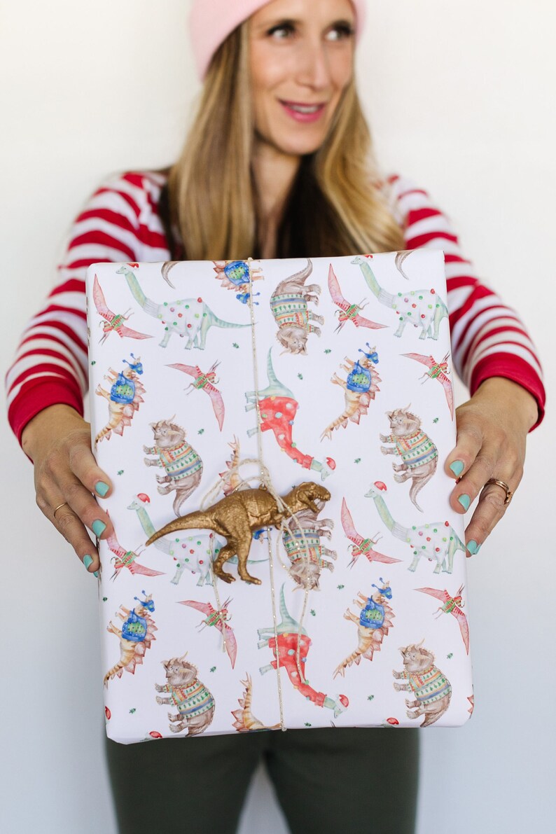 May include: A white gift wrap with a repeating pattern of dinosaurs wearing sweaters. A gold dinosaur toy is attached to the wrapping paper with twine.