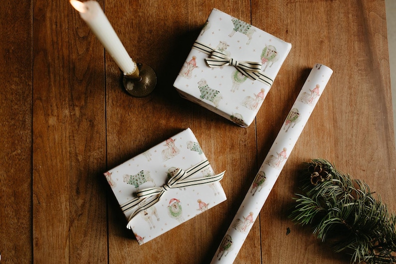 May include: Two wrapped gifts with striped bows and a roll of matching wrapping paper featuring a festive llama design. A lit candle and pine sprig with a pinecone are on a wooden surface.