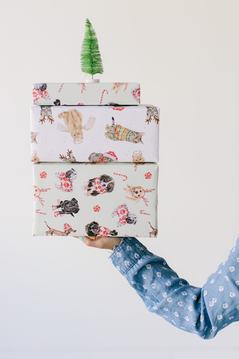 Puede incluir: Un conjunto de tres rollos de papel de regalo con un tema navide&ntilde;o festivo. El rollo superior presenta un &aacute;rbol de Navidad verde, el rollo del medio presenta gatos con gorros de Pap&aacute; Noel y su&eacute;teres, y el rollo inferior presenta perros con gorros de Pap&aacute; Noel y su&eacute;teres.