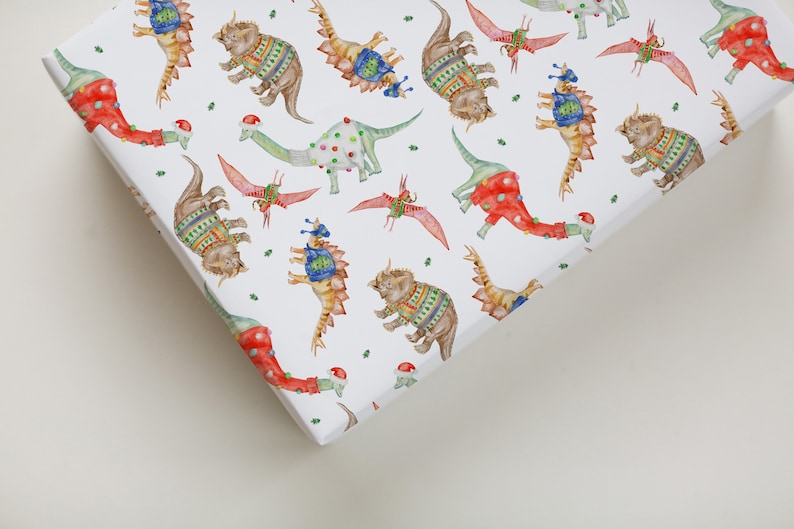 May include: White wrapping paper with a repeating pattern of dinosaurs wearing festive sweaters and hats. The dinosaurs are in various colors, including red, green, blue, and yellow.