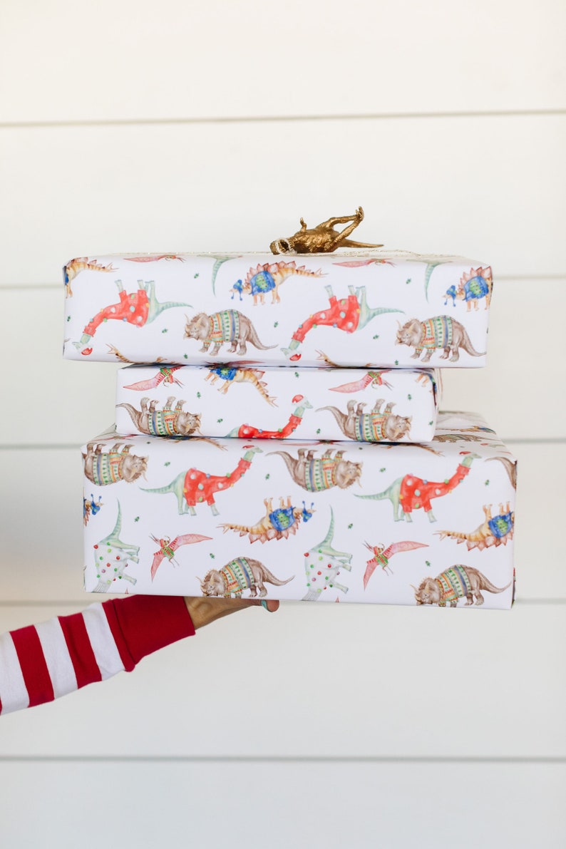 May include: Three wrapped gifts with a dinosaur pattern. The wrapping paper is white with red, blue, and green dinosaurs wearing sweaters. The gifts are stacked on top of each other.