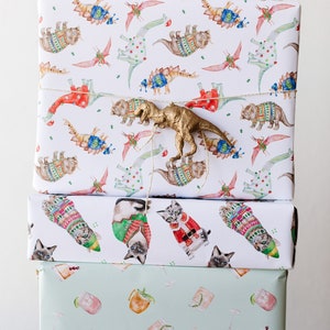 May include: Three wrapped gifts stacked on top of each other. The top gift is wrapped in white paper with a repeating pattern of dinosaurs wearing sweaters. A gold plastic dinosaur is on top of the gift. The middle gift is wrapped in white paper with a repeating pattern of cats wearing sweaters. The bottom gift is wrapped in light green paper with a repeating pattern of cocktails.