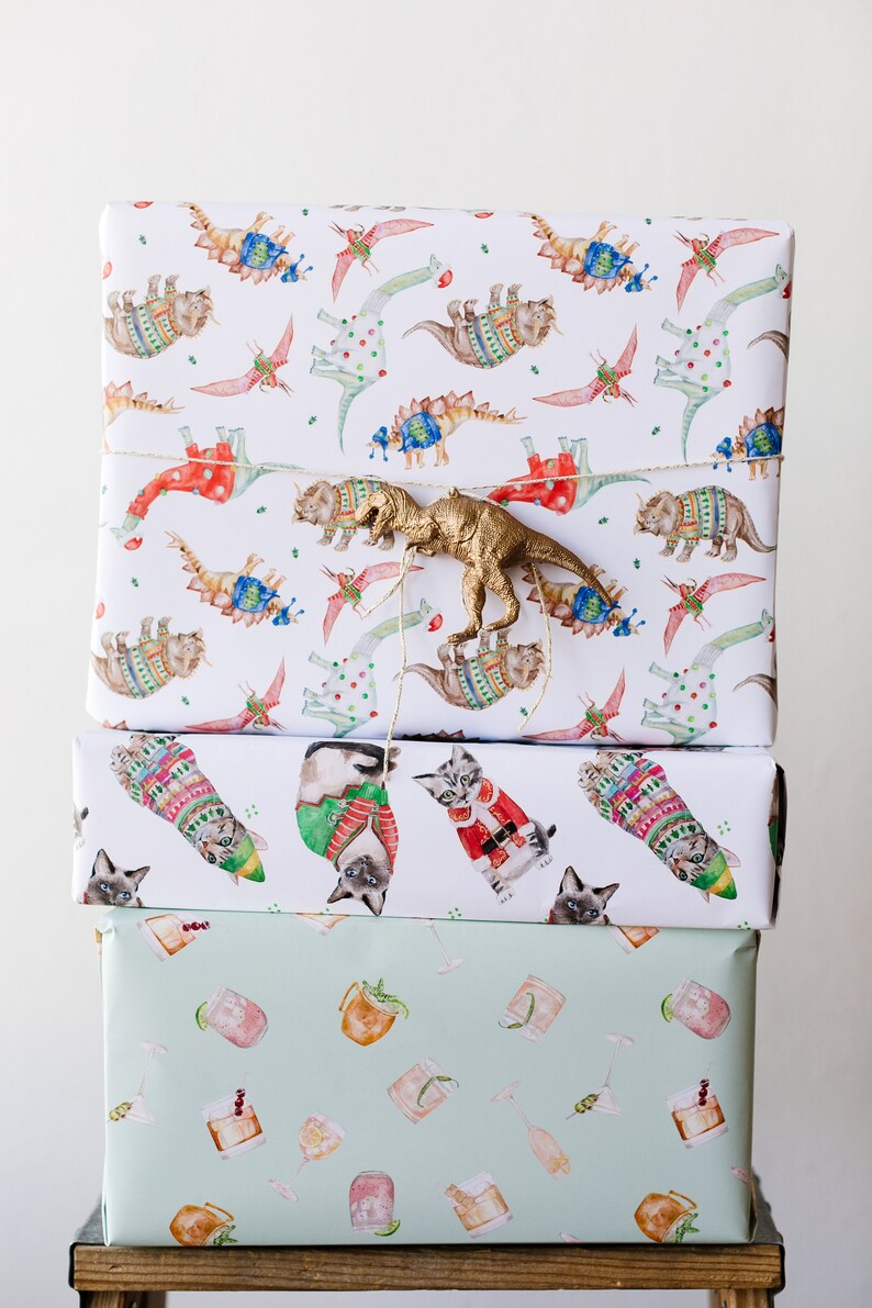 May include: Three wrapped gifts stacked on top of each other. The top gift is wrapped in white paper with a repeating pattern of dinosaurs wearing sweaters. A gold plastic dinosaur is on top of the gift. The middle gift is wrapped in white paper with a repeating pattern of cats wearing sweaters. The bottom gift is wrapped in light green paper with a repeating pattern of cocktails.