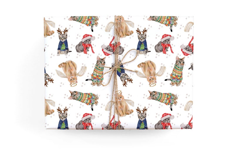 Puede incluir: Papel de regalo blanco con un patr&oacute;n repetitivo de gatos con su&eacute;teres navide&ntilde;os, cuernos de reno y alas de &aacute;ngel. Los gatos est&aacute;n ilustrados en estilo acuarela.