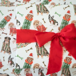 May include: A white gift wrap with a red ribbon and bow. The wrapping paper features a repeating pattern of dogs wearing festive plaid outfits and Santa hats. The dogs are surrounded by scattered bone-shaped designs.