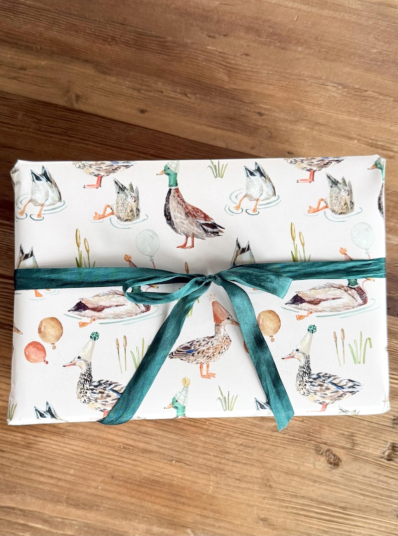 May include: White gift wrap featuring a pattern of ducks wearing party hats and holding balloons. The wrap is tied with a green ribbon.