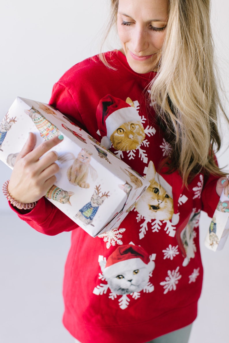 Puede incluir: Una mujer lleva un su&eacute;ter rojo de Navidad con un patr&oacute;n de gato. El su&eacute;ter tiene copos de nieve y gatos con gorros de Pap&aacute; Noel. Ella sostiene un regalo envuelto en papel blanco con un patr&oacute;n de gato.