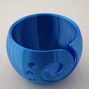 Könnte beinhalten: Eine blaue Garnschale mit kugelförmiger Form und einem gebogenen Schlitz für Garn. Die Schale hat drei kleine Löcher und ein dekoratives Wirbeldesign an der Seite. Dieser Artikel ist für Strick- und Häkelfans konzipiert.