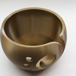 Puede incluir: Un cuenco de hilo de madera marrón dorado con una forma lisa y redondeada. El cuenco tiene una ranura curva para el hilo y varios agujeros en el lateral. Este cuenco artesanal está diseñado para proyectos de punto y ganchillo.