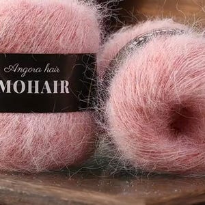 Może przedstawiać: Dwa puszyste, różowe motki wełny. Jeden ma czarną etykietę z napisem "Angora hair MOHAIR". Wełna ma miękką, lekko puszystą fakturę, idealną do projektów dziewiarskich lub szydełkowych. Wełna jest prezentowana na drewnianej powierzchni.
