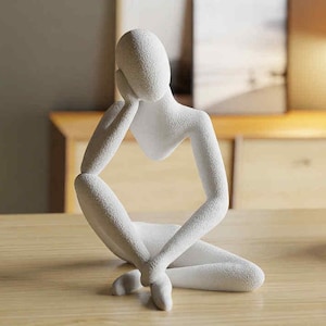 Könnte beinhalten: Eine weiße, strukturierte Skulptur einer Person in sitzender, kontemplativer Pose. Die Figur stützt den Kopf auf eine Hand, die Beine sind gekreuzt. Die Skulptur steht auf einer Holzoberfläche, im Hintergrund Kerzen und eine Lampe.