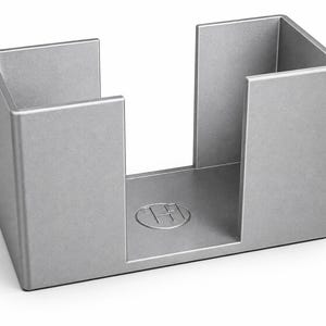 Könnte beinhalten: Ein silberfarbener Serviettenhalter aus Metall mit U-förmigem Design und offenen Seiten. Die Basis zeigt ein kreisförmiges, geprägtes Logo mit den Buchstaben TH im Inneren. Die Struktur hat eine glatte, matte Oberfläche und eine rechteckige Form.