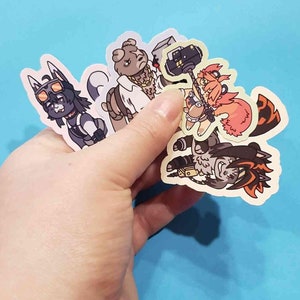 Puede incluir: Una mano sosteniendo una colección de pegatinas de personajes de dibujos animados coloridos. Las pegatinas presentan varios animales antropomórficos con diseños únicos, incluyendo un perro con gafas y un oso con una cadena de oro.