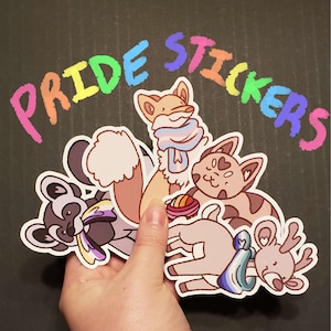 May include: A collection of Pride stickers featuring various animal illustrations, including a fox, raccoon, cat, and deer. The stickers have a white border and are held against a dark background. The words "Pride Stickers" are written in rainbow colors.