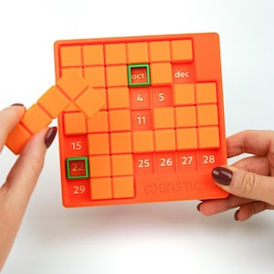 May include: An orange 3D calendar puzzle with a grid of squares. The calendar has the months "oct" and "dec" printed on it. A hand is placing an orange block into the grid. The numbers 4, 5, 11, 15, 22, 25, 26, 27, 28, and 29 are visible.