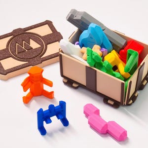 May include: A wooden treasure chest filled with colourful 3D printed puzzle pieces. The chest has a brown lid with a logo of a mountain range. There are four additional puzzle pieces outside of the chest, including a bright orange figure, a blue piece, a pink piece, and a blue piece.