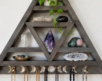 May include: A triangular dark gray wooden wall shelf with multiple horizontal tiers. The shelf displays a potted green plant, various crystals, a small bowl, and a geode. The bottom section features silver moon phase icons and hooks holding several hanging necklaces.