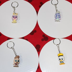 May include: Four character keychains on white circles against a red background with Mickey Mouse silhouettes. The keychains feature cartoon characters with silver key rings. The characters are in various colors, including yellow, blue, and brown.