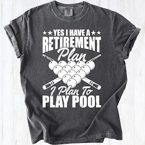 May include: Dark gray t-shirt with a white graphic that reads "YES I HAVE A RETIREMENT Plan I Plan To PLAY POOL". The design includes pool balls and crossed pool cues. The shirt has rolled-up sleeves.