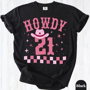 May include: A black short-sleeve t-shirt featuring the pink text HOWDY above a pink cowboy hat and the number 21. The design includes pink star accents and a pink and light pink checkered pattern along the bottom edge of the graphic.